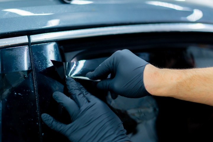 Close-up of a technician wearing black gloves applying and smoothing window tint film on a car window using a tool